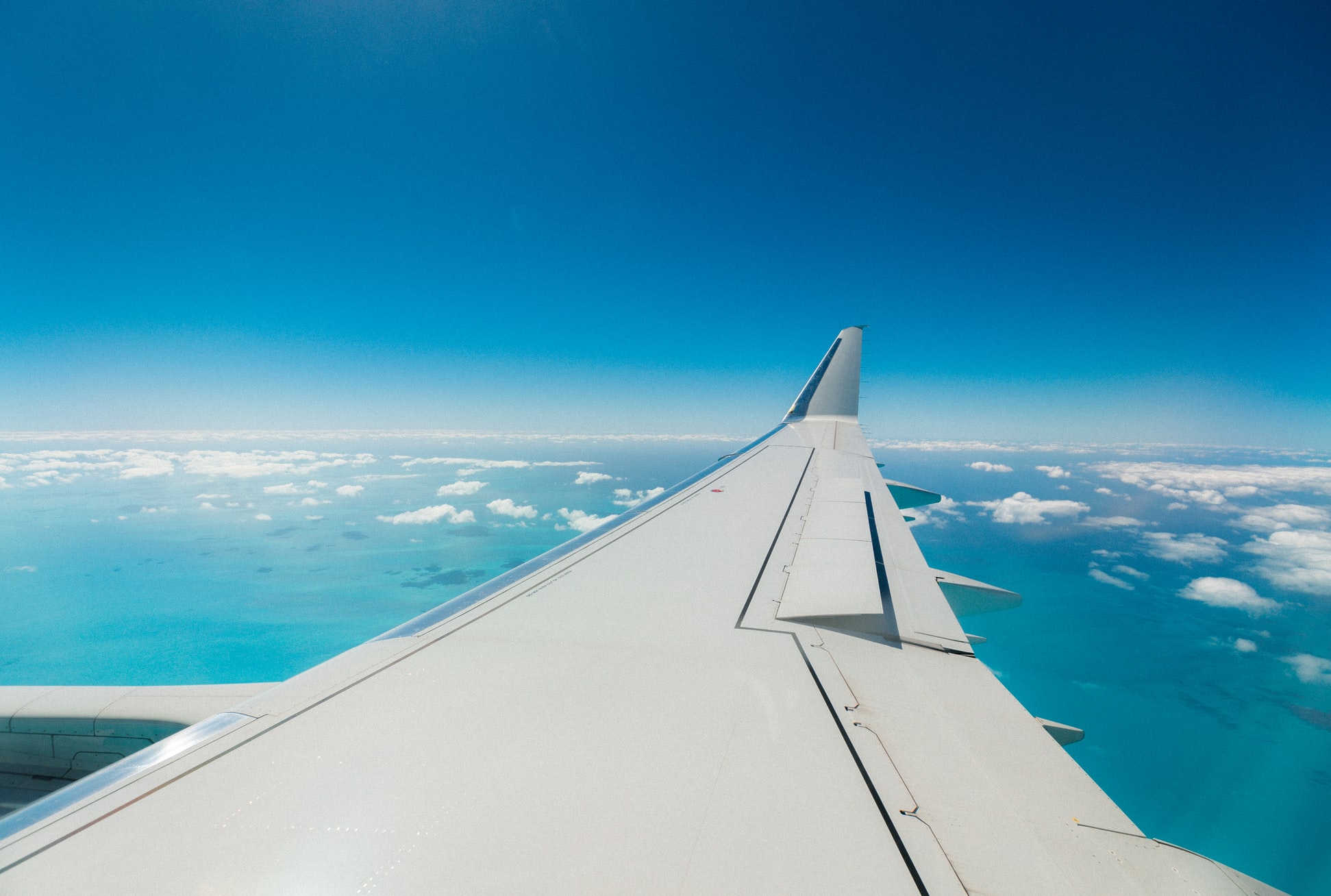View from an airplane window of a white wing with winglet, above clouds and turquoise ocean water, under a deep blue sky